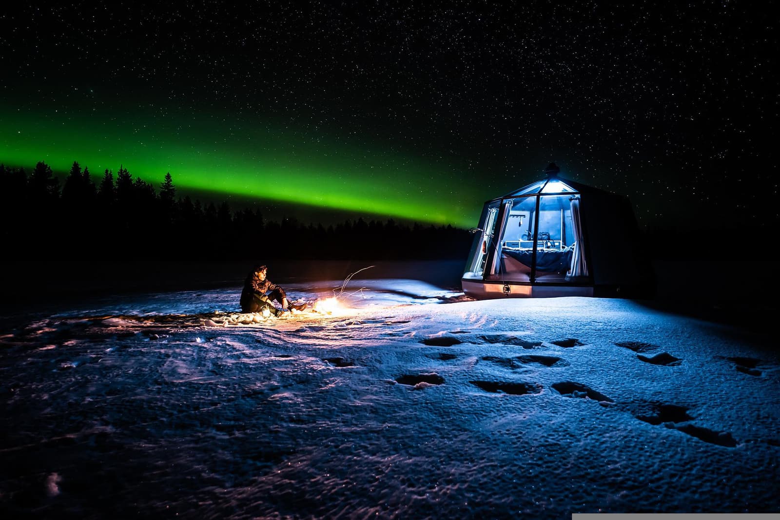 A glass igloo and a campfire on the snow beneath a green band of northern lights in Finnish Lapland