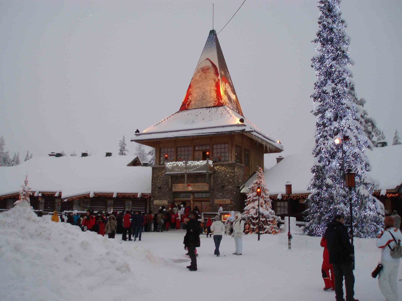 The snow-covered main building of Santa Claus Village with its pointed roof, and visitors in the square