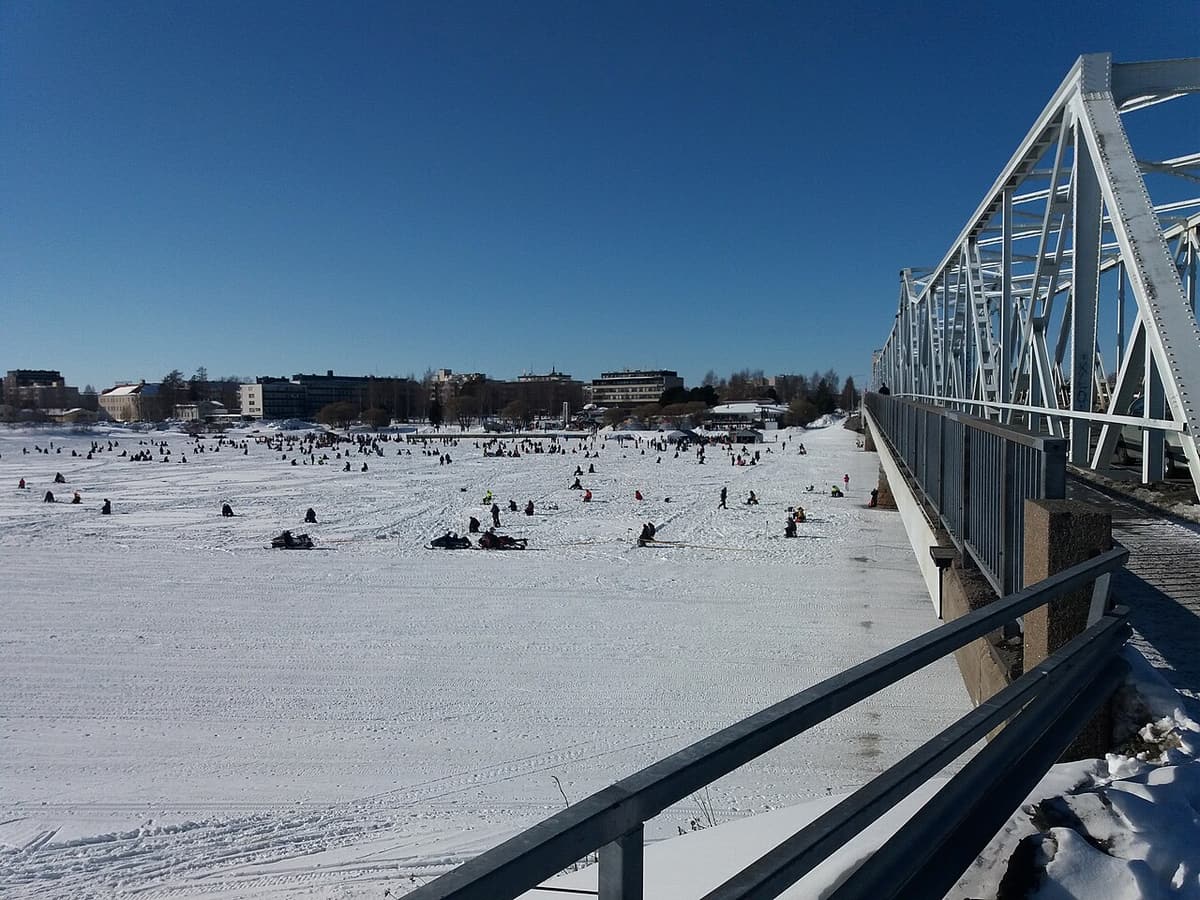 Ice fishers scattered across a wide frozen river beside a steel bridge in northern Finland