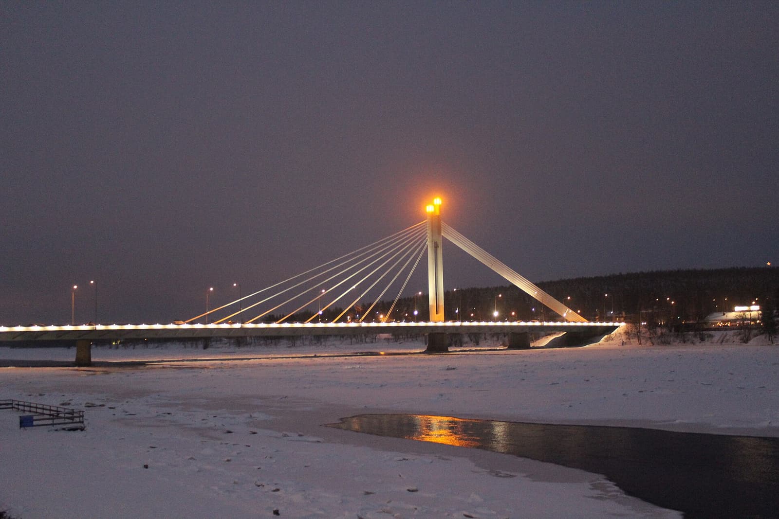 The Lumberjack’s Candle Bridge lit up at night above the snowy riverbank