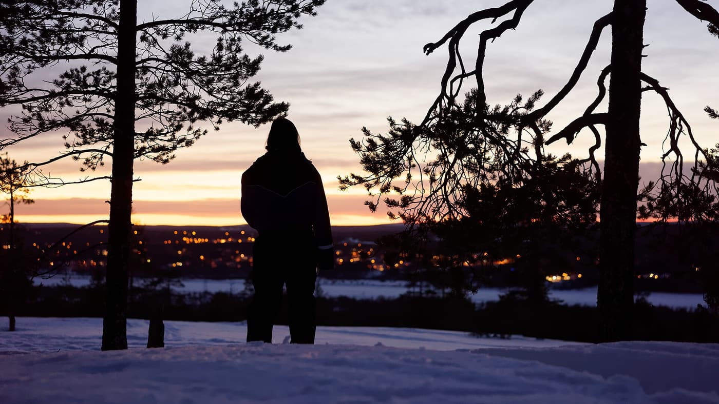 A lone figure silhouetted among pines at dusk above the lights of Rovaniemi