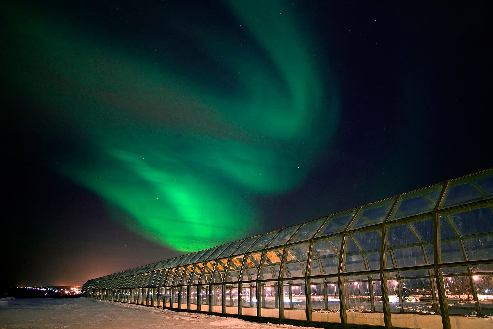 A bright green aurora curling over the lit glass corridor of the Arktikum museum in Rovaniemi